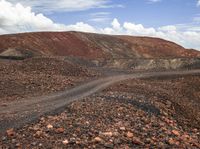 a dirt and rock road with trees in the distance near a hill with mountains on one side