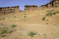 a dirt road in front of rocky cliffs in a desert area with plants and weeds