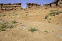 a dirt road in front of rocky cliffs in a desert area with plants and weeds
