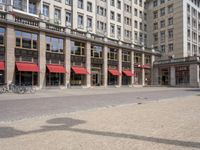 two people sit on a bench in front of a building with many windows and a red awning