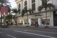 people walking on the side walk next to a building and palm trees in front of a store