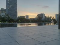 the walkway at the park leads to a lake and buildings in the background as evening falls