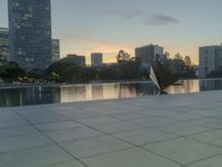 the walkway at the park leads to a lake and buildings in the background as evening falls