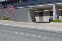 a car is parked in front of an office building on a street corner with a concrete parking garage and flowering trees on the sidewalk