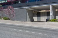 a car is parked in front of an office building on a street corner with a concrete parking garage and flowering trees on the sidewalk