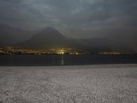 a bench sitting near the ocean during night with lights on mountains in the background on a cloudy day