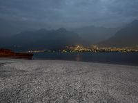 a bench sitting near the ocean during night with lights on mountains in the background on a cloudy day