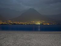a bench sitting near the ocean during night with lights on mountains in the background on a cloudy day