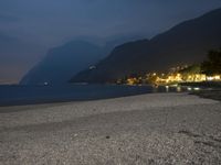 a bench sitting near the ocean during night with lights on mountains in the background on a cloudy day