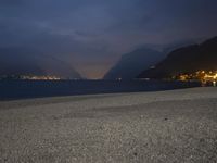 a bench sitting near the ocean during night with lights on mountains in the background on a cloudy day