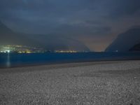 a bench sitting near the ocean during night with lights on mountains in the background on a cloudy day