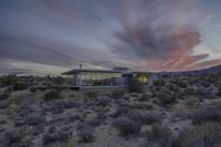 a house in the desert lit up at dusk with clouds overhead and mountains in the background