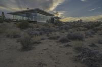 a modern home sits in the desert on a cloudy day at dusk with clouds over it