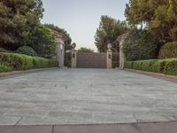 the driveway entrance leads to a gate and outdoor privacy paneled doors, which are lined with greenery