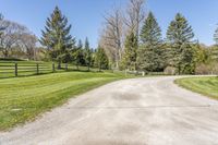 a paved driveway leading into a tree lined field with lots of green grass and trees