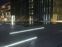 several light strips run along the sidewalk with buildings in the background at night time in the city