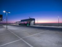 a parking lot next to the ocean at dusk, with empty vehicles parked in the street