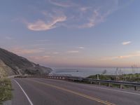 a highway with sea in the distance at dusk and cliffs above, and a hill behind it