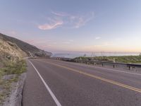 a highway with sea in the distance at dusk and cliffs above, and a hill behind it