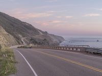 a highway with sea in the distance at dusk and cliffs above, and a hill behind it