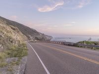 a highway with sea in the distance at dusk and cliffs above, and a hill behind it