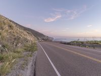 a highway with sea in the distance at dusk and cliffs above, and a hill behind it