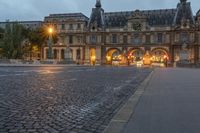 a street and the sky at dusk near a large building with an arch in front