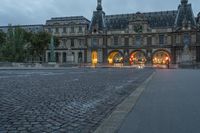 a street and the sky at dusk near a large building with an arch in front