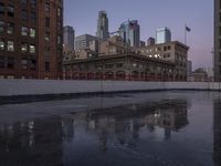 a puddle on a road is reflecting some buildings in the water at dusk by buildings