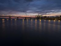 a long bridge spanning across a large body of water at dusk with lights shining on the side