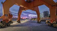 a view from under an orange crane at dusk near a large city skyline, with construction on the banks