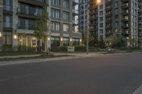 an empty street lined with tall buildings at dusk over a green field with some grass