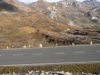 the asphalt road is lined with rocks in front of the mountains, surrounded by a grassy hillside