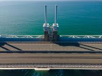 a bridge over the ocean with construction equipment in the background and two people walking on it