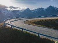 the sun is shining brightly on a curved road with mountains in the distance, with snow capped peaks