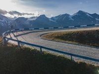 the sun is shining brightly on a curved road with mountains in the distance, with snow capped peaks