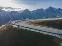 the sun is shining brightly on a curved road with mountains in the distance, with snow capped peaks