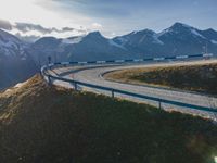 the sun is shining brightly on a curved road with mountains in the distance, with snow capped peaks