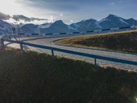 the sun is shining brightly on a curved road with mountains in the distance, with snow capped peaks