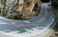 a motorcycle is going down an empty mountain road under some trees and rocks, turning to a curve into a bend