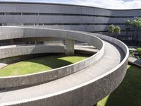 a circular green lawn and concrete walkway with a round shaped building and trees in the background