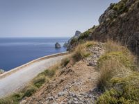 a car drives down the road alongside a rocky cliff on a beach side with blue sea and sky in the background