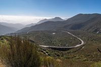 a curved road on the side of mountains with bushes on both sides and hills with clouds overhead