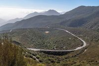 a curved road on the side of mountains with bushes on both sides and hills with clouds overhead