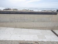 a person walking up a sidewalk next to the ocean and cement wall with waves in background