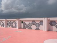 an empty basketball court with painted concrete and pink concrete walls in front of storm clouds