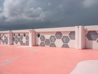 an empty basketball court with painted concrete and pink concrete walls in front of storm clouds