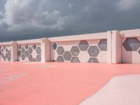 an empty basketball court with painted concrete and pink concrete walls in front of storm clouds