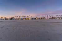 an empty beach with sand, palm trees, and sunset lighting in the distance is a beach, palm trees in the foreground, sand, there are houses on the beach area with a small footprints,