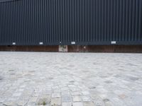 an empty bench sitting on top of a brick pavement in front of a black wall